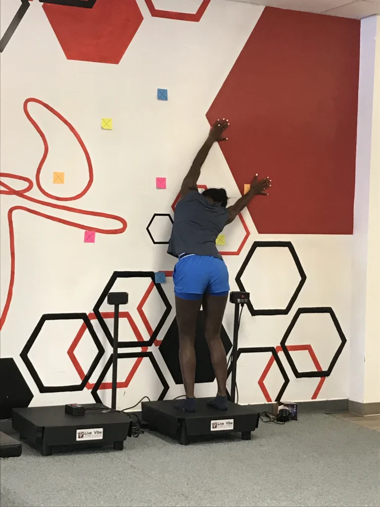 Woman standing on vibration machine stretching against the wall.